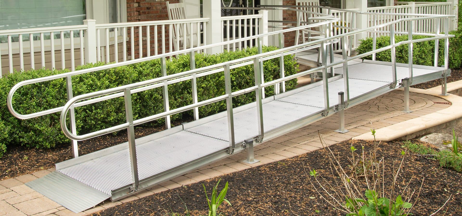 Modular aluminum ramp with handrails running alongside a white front porch and boxwood hedges
