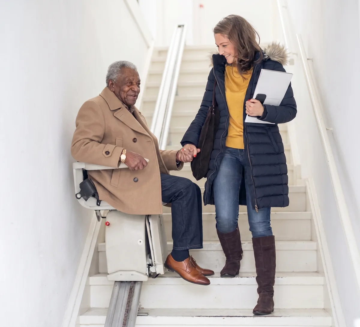 An older man riding a stairlift down a bright staircase, holding hands with a woman walking down the steps beside him